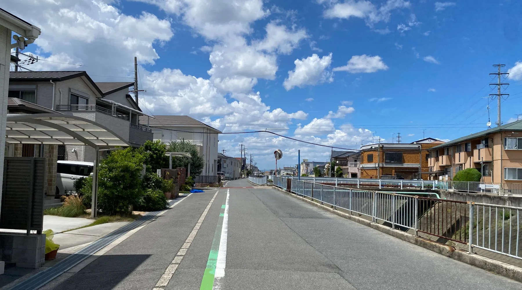 白い雲が浮かぶ青空の下、住宅が並ぶまちの通りと水路沿いの柵
